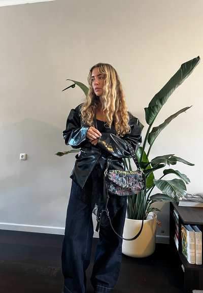 Mujer con cabello largo y rizado, vestida con una chaqueta negra brillante y pantalones anchos, sosteniendo un bolso colorido con estilo graffiti, junto a una gran planta en maceta.