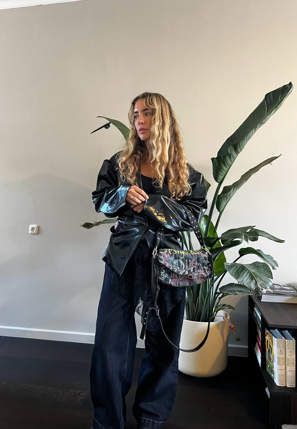 Femme aux longs cheveux bouclés portant une veste noire brillante et un pantalon ample, tenant un sac à main coloré au style graffiti, debout auprès d'une grande plante en pot.