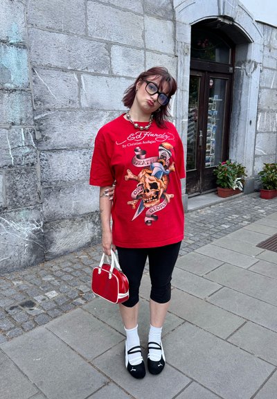 Camiseta roja de gran tamaño con gráfico de calavera, leggins negros, calcetines blancos, zapatos negros y un pequeño bolso rojo. Fondo de pared de piedra.