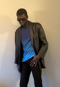 Long manteau en cuir noir sur une chemise dégradée bleue, pantalon noir et lunettes de soleil. Bague remarquable à la main gauche, debout contre un mur clair.