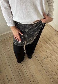 Personne portant un pantalon noir et un haut blanc à manches longues ajustant un tablier en dentelle noire transparente autour de sa taille sur un sol en bois.