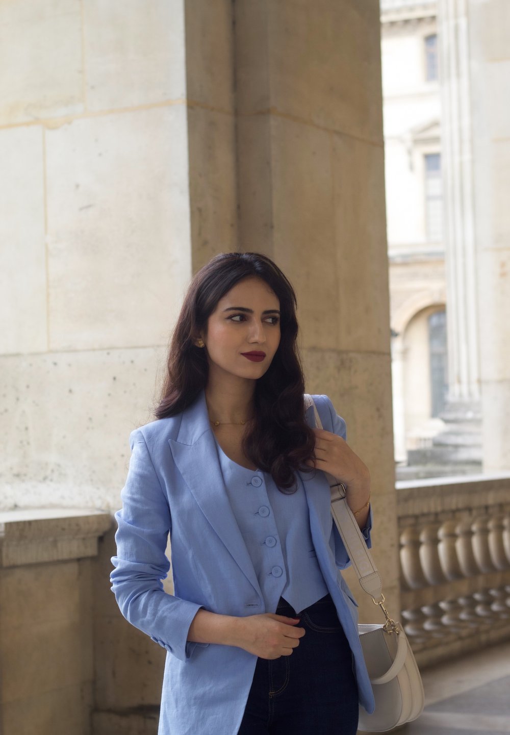 Lyseblå blazer over en matchende knaptrøje, parret med mørke jeans. En hvid håndtaske hænger på den ene skulder; glat, struktureret stof.