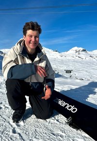 Joven arrodillado sobre la nieve junto a una tabla de snowboard con la etiqueta "Zalando", con montañas nevadas y un cielo azul claro de fondo.