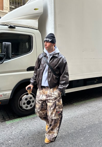 Joven con gorro, chaqueta de cuero, pantalones cargo de camuflaje y zapatos amarillos sosteniendo un café, de pie junto a un gran camión de reparto blanco en la calle.