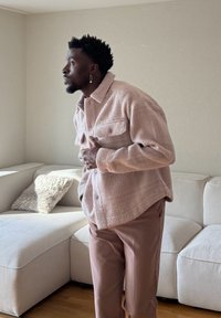 Homme debout à l'intérieur, portant une veste texturée rose clair et un pantalon marron, avec des boucles d'oreilles, regardant vers la gauche près d'un canapé blanc.