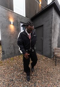 Homme aux cheveux tressés portant une veste et un pantalon noirs, debout sur un sol rocheux près d'un bâtiment gris avec une porte et un banc.