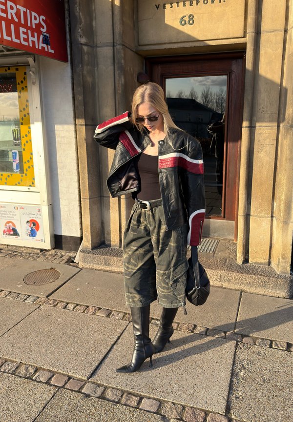 Mladá žena v slnečných okuliaroch, v čiernej koženej bunde s červenými pruhmi, maskáčových cargo nohaviciach a čiernych členkových čižmách na opätku stojí na chodníku pri kamennom vchode do budovy.