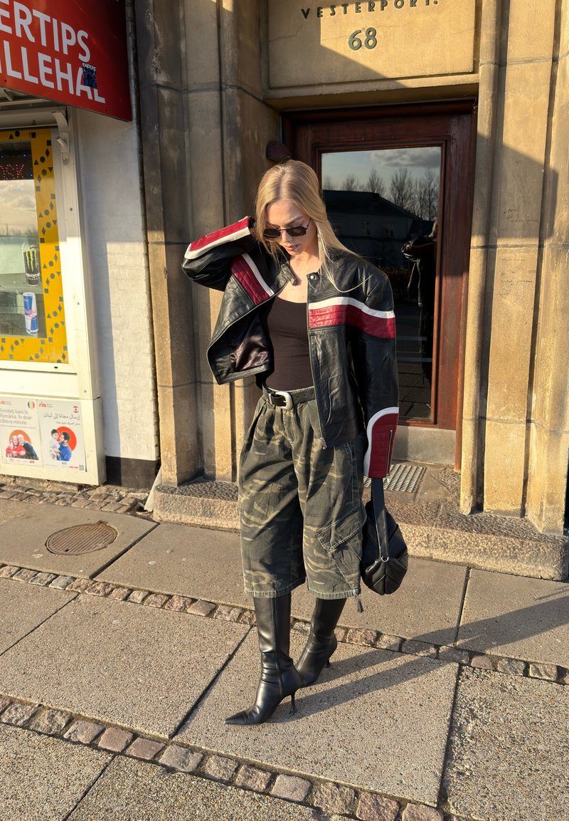 Jeune femme portant des lunettes de soleil, une veste en cuir noire avec des bandes rouges, un pantalon cargo camouflage et des bottes noires à talons, debout sur le trottoir près de l'entrée d'un bâtiment en pierre.