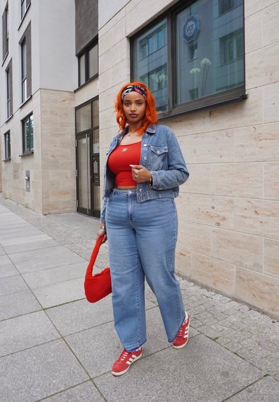 Chaqueta de mezclilla, top rojo de tirantes, pantalones anchos de mezclilla azul claros, bolso de punto rojo y zapatillas rojas con acentos blancos. Entorno urbano con paredes de piedra.