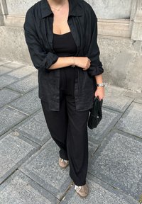 Kobieta w czarnej bluzce, czarnej koszuli zapinanej na guziki i czarnych spodniach, trzymaj�ąca czarną plecioną torebkę na kamiennej nawierzchni w pobliżu ściany budynku.