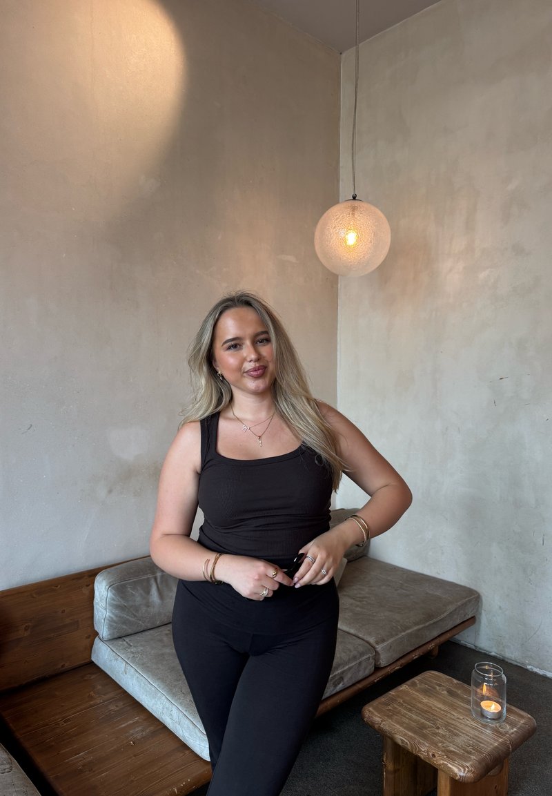 Schwarzes, geripptes Tanktop kombiniert mit schwarzen Leggings, vor einer strukturierten beigen Wand, daneben eine Glas-Pendelleuchte und Holzmöbel.