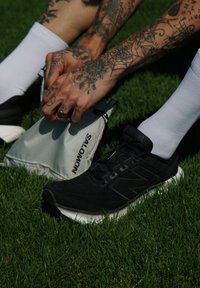 Zapatilla deportiva negra con una parte superior texturizada y suela blanca, combinada con calcetines blancos; una pequeña bolsa gris de Salomon sobre césped verde.