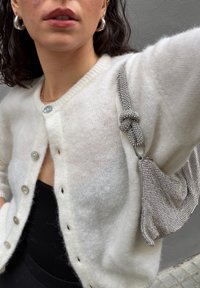 Valge, pehme kardigan ümmarguste nööpidega, mille kõrvale on komplekteeritud hõbedane metallik käekott sõlmitud rihma ja tekstuurse pinnaga.