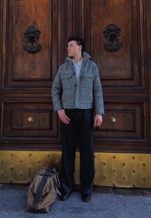 Hombre con chaqueta a cuadros gris y pantalones negros está junto a una gran puerta de madera con aldabas decorativas, al lado de una bolsa de viaje marrón en el suelo.