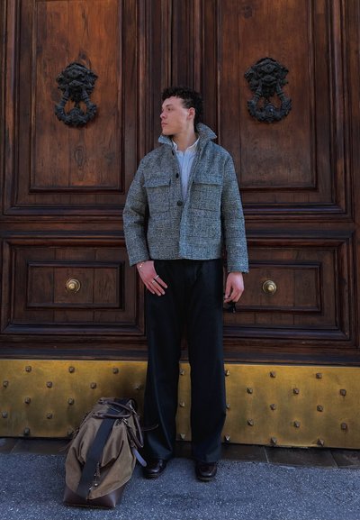 Hombre con chaqueta a cuadros gris y pantalones negros está junto a una gran puerta de madera con aldabas decorativas, al lado de una bolsa de viaje marrón en el suelo.