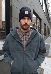 Pilkas užtraukiamas džemperis su užrašu „VERVER“ ir „BERLIN“, dėvimas ant rudos languotos marškinių. Derinamas su juoda kepure ir tamsiomis džinsinėmis kelnėmis.