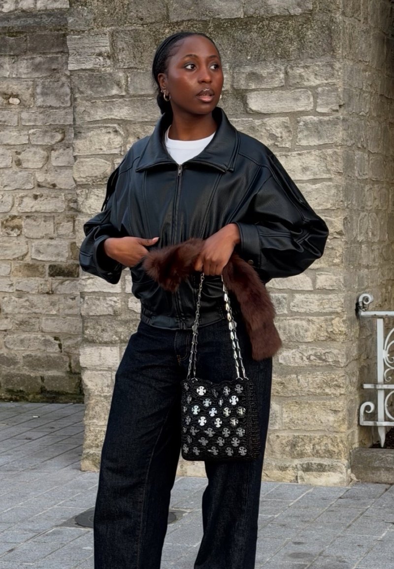 Femme portant une veste en cuir noire et un pantalon foncé, tenant un sac à main noir à motifs avec un accessoire en fourrure marron, debout près d'un mur en pierre.
