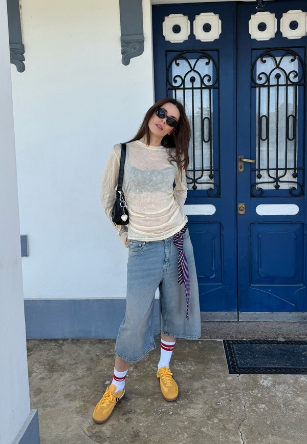 Donna con occhiali da sole, maglietta trasparente a maniche lunghe, pantaloni larghi in denim, scarpe gialle e calzini bianchi davanti a una porta doppia blu.