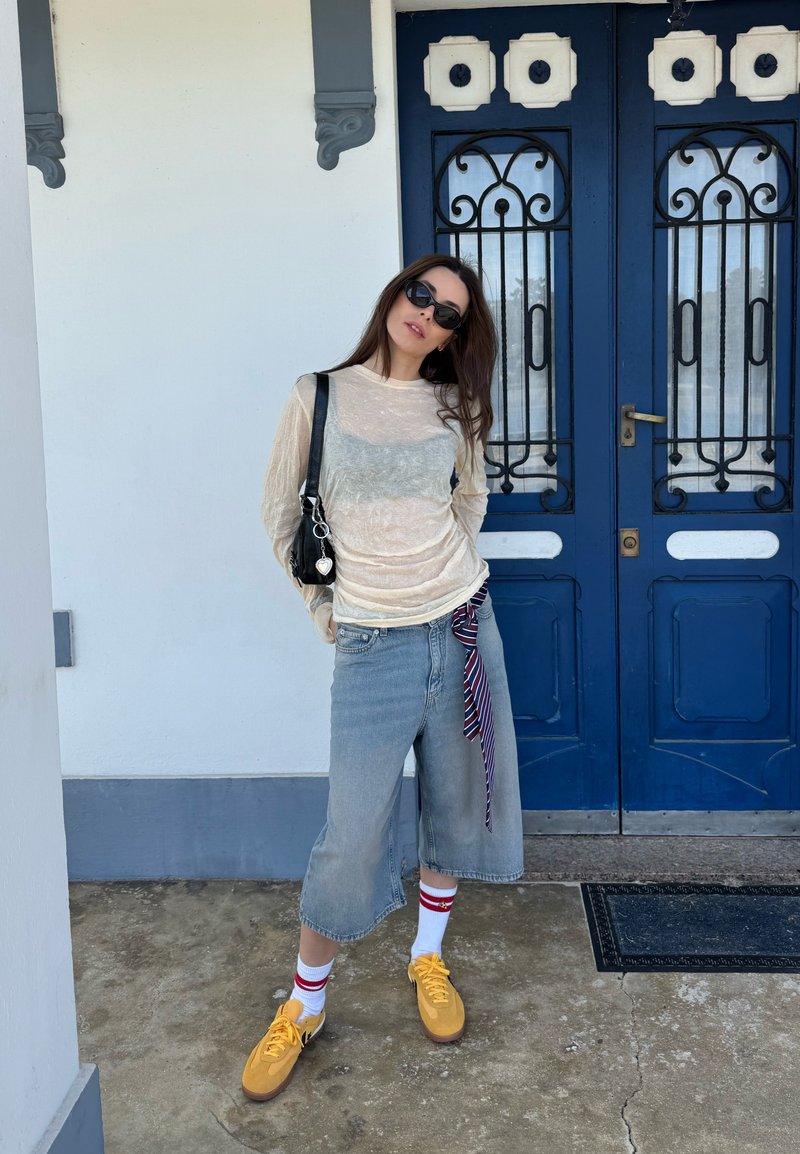 Femme portant des lunettes de soleil, un haut transparent à manches longues, un pantalon large en denim, des chaussures jaunes et des chaussettes blanches debout devant des portes doubles bleues.