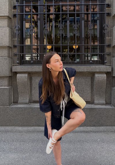 Joven mujer con cabello largo, con chaqueta vaquera oscura y pantalones cortos, ajusta un zapato blanco, de pie sobre pavimento gris junto a un edificio de piedra con ventana con rejas.
