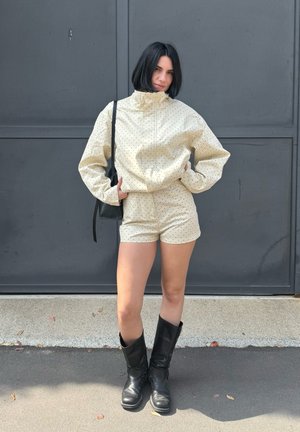 Mujer con cabello bob negro, que lleva una chaqueta beige con lunares y pantalones cortos, botas negras y un bolso de hombro negro, de pie contra una pared oscura.