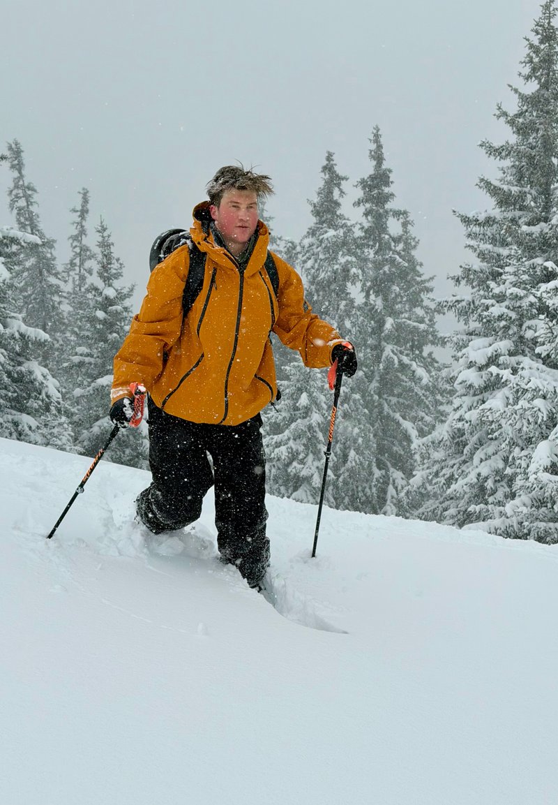 Personne en veste orange faisant de la randonnée dans une neige profonde avec des bâtons de trekking dans une forêt enneigée avec des conifères couverts de neige.