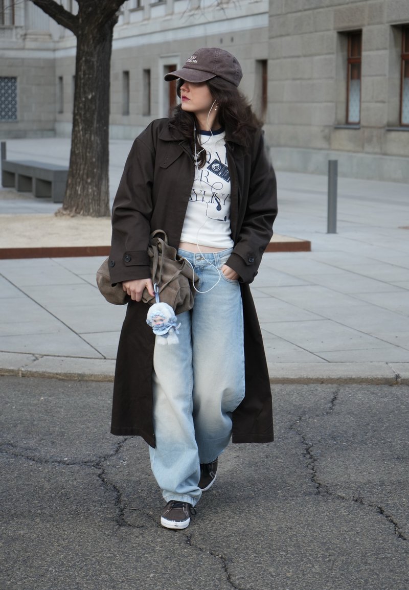 Femme portant un long manteau sombre, une casquette de baseball, un jean ample et des baskets, marchant dans une rue de la ville, tenant un sac avec un porte-clés en peluche et des écouteurs dans les oreilles.
