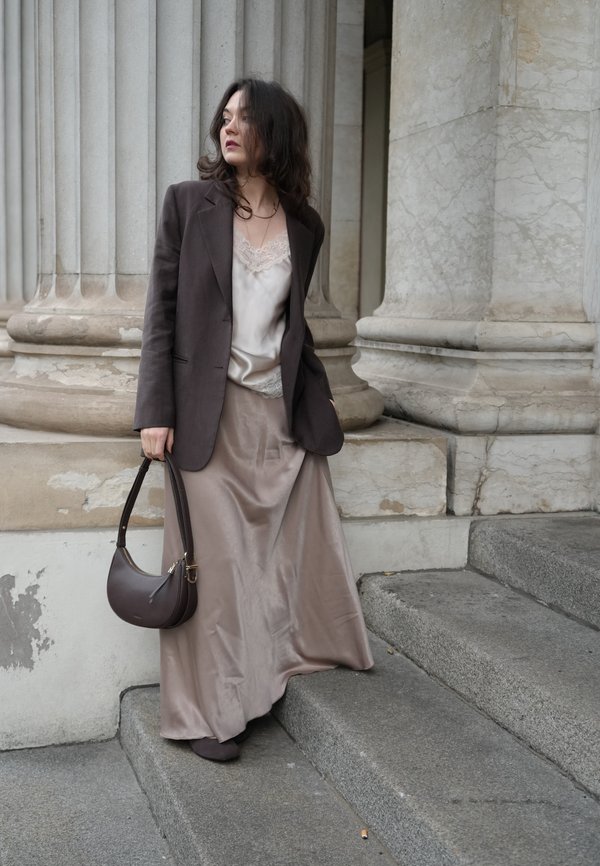 Blazer marron sur mesure porté sur un haut en satin léger orné de dentelle, assorti à une jupe fluide en satin beige et un sac à main marron. Des marches en arrière-plan.