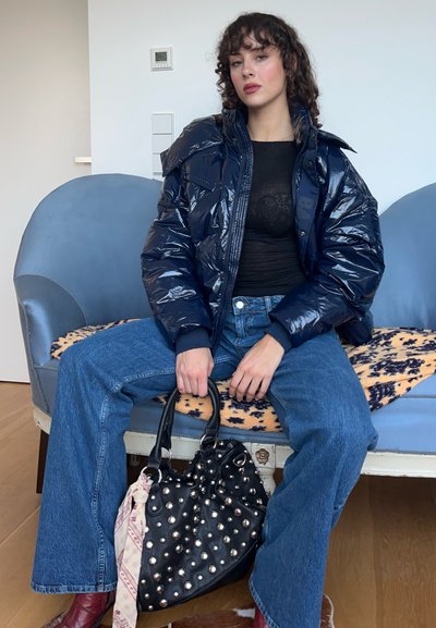 Chaqueta de plumas azul marino, blusa de encaje negra, jeans de denim azul y un bolso negro con tachuelas y una bufanda estampada. Cabello rizado, en pose sentada.