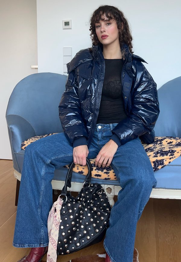 Marine pufferjack, zwarte kanten top, blauwe denim jeans en een zwarte studded handtas met een patroon sjaal. Krullend haar, zittende pose.