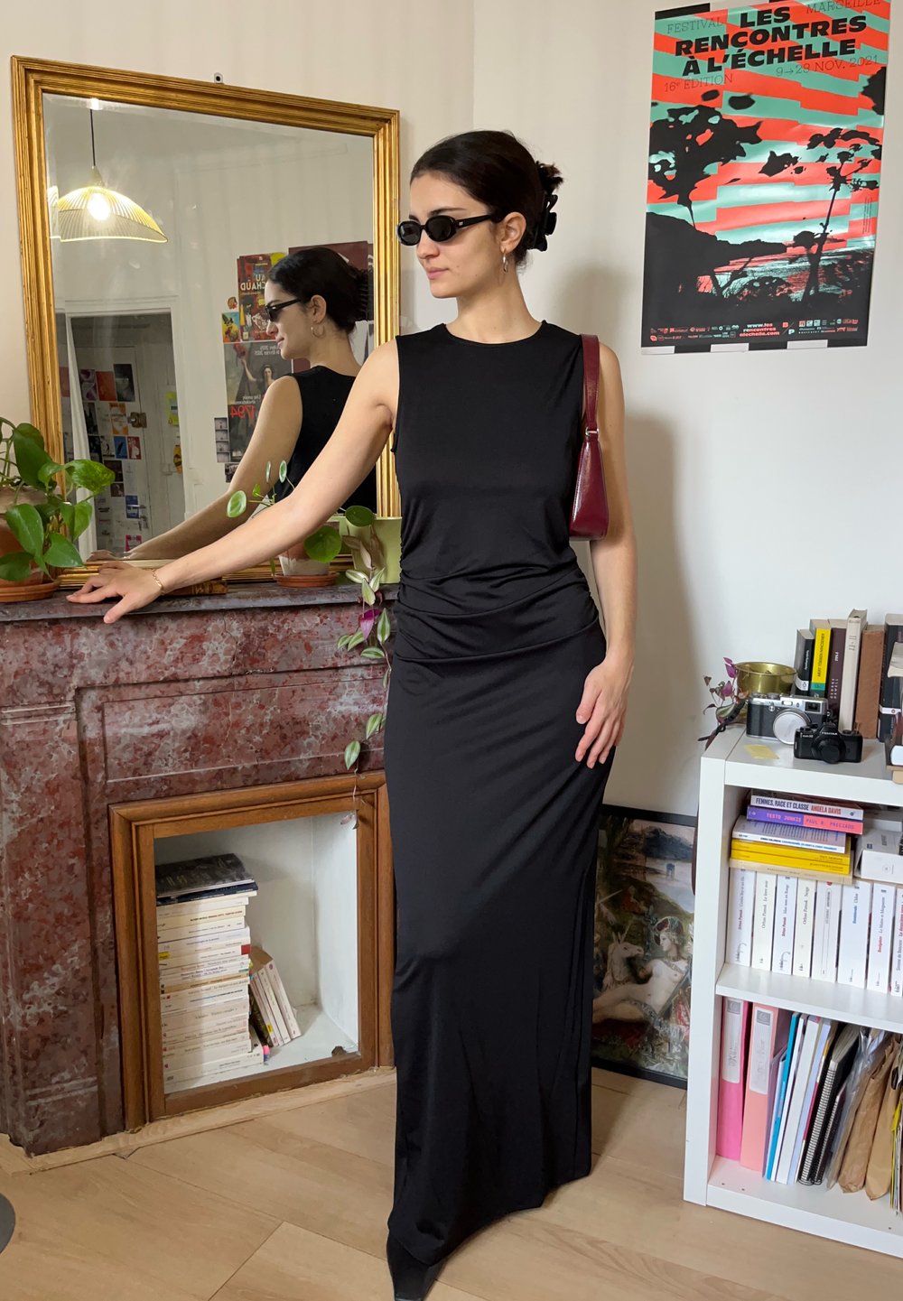 Femme en robe noire sans manches et lunettes de soleil, debout près d'une cheminée en marbre avec des plantes et un miroir, à côté d'une bibliothèque avec des livres et des appareils photo.