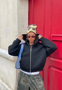 Sweat à capuche zippé noir, bonnet à motifs avec accents jaunes, pantalon camouflage et sac bleu avec clous argentés. Fond mural rouge.