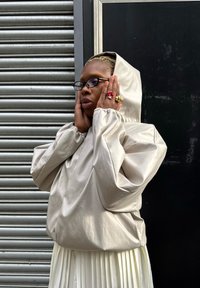 Lichtbeige capuchonjack met losse pasvorm, elastische manchetten en een gladde textuur, gecombineerd met een witte geplooide rok. Opvallende ringen aan de handen.