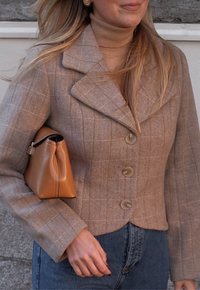 Blazer beige à motif pied-de-poule texturé avec un col cranté et trois boutons, associé à un sac à main en cuir beige sur un fond clair.