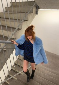 Mlada ženska z rdečimi lasmi, oblečena v modri krzneni plašč, črno-modro črtast pulover, kratko krilo in črne škornje, ki se povzpela po betonskih stopnicah.