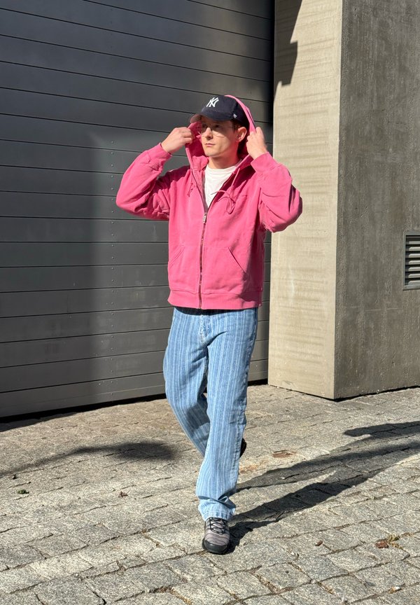 Eine junge Person richtet einen rosa Kapuzenpullover, während sie auf einem Kopfsteinpflaster in der Nähe einer grauen Wand und einer Beton Säule im Sonnenlicht geht.
