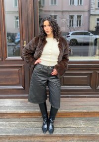 Femme aux cheveux bouclés portant une veste en fourrure marron, un haut blanc boutonné, un short en cuir noir et des bottes noires montantes, debout sur des marches en bois.