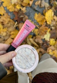 Baume à lèvres hydra-peptide de Nudestix dans un tube rose et illuminateur Revolution dans une pochette transparente, présentant une texture tourbillonnante et scintillante.