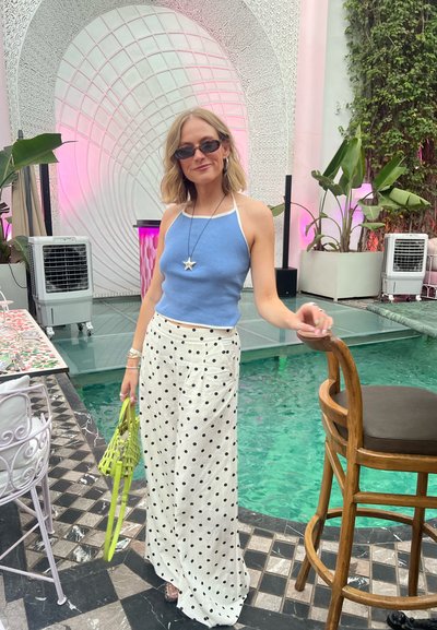 Top halter azul claro, falda blanca con lunares negros, bolso verde, cabello corto y rizado, de pie junto a una piscina de azulejos con plantas.