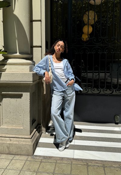 Camisa a rayas azul y blanca, top blanco sin mangas, jeans de pierna ancha azul claro y zapatos blancos. El escenario presenta una columna de piedra texturizada y un suelo con patrón.