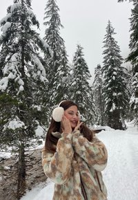 Beige Kunstfelljacke mit grünen und braunen Mustern, mit einem Reißverschluss. Weiße, flauschige Ohrenschützer. Schneeiger Wald Hintergrund.