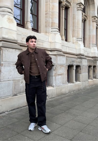 Joven con chaqueta marrón, pantalones oscuros y zapatillas blancas de pie en la acera de la ciudad junto a un edificio de piedra con ventanas altas.