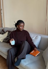 Pull tricoté marron avec manches à franges, pantalon foncé, tenant une tasse bleue texturée, assis sur un canapé gris clair, près d'un livre orange.