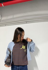 Femme portant un cardigan bleu par-dessus une chemise à rayures marron avec un motif jaune, tenant un sac noir, debout devant un mur blanc avec des tuyaux au-dessus.