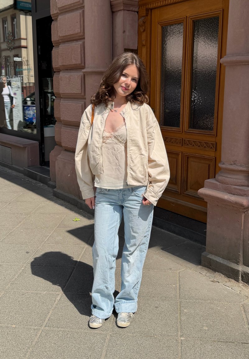 Jeune femme aux cheveux bruns portant une veste crème, un top en dentelle et un jean bleu clair, debout sur un trottoir ensoleillé près d'une porte en bois.