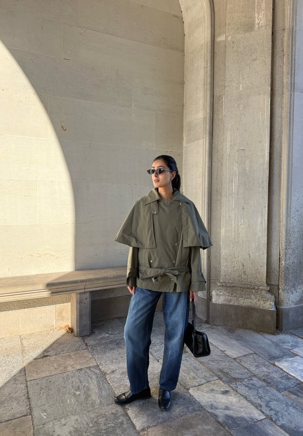 Una mujer con gafas de sol, una chaqueta verde oliva estilo capa, vaqueros y zapatos negros está de pie junto a un muro de piedra y lleva un bolso negro.