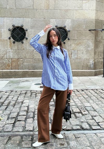 Camisa de botones a rayas azul y blanca con un cuello blanco, combinada con pantalones anchos marrones y zapatos blancos, sosteniendo un bolso negro.