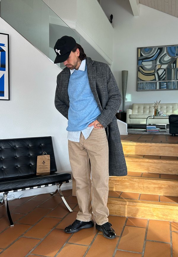 Uomo con berretto nero, cappotto grigio a quadri, maglione blu, pantaloni beige e scarpe nere in piedi all'interno vicino a scale e panca in pelle nera con una borsa di carta.