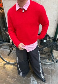 Maglione rosso di lana sopra una camicia rosa chiaro a righe, abbinato a pantaloni neri larghi con strisce laterali bianche. Anello d'argento e bicicletta sullo sfondo.