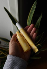 Twee huidverzorgingsproducten in gele verpakking vastgehouden in een hand. Een druppelflesje met het label "Vitamine Elixir" en een tube met het label "Collageen."